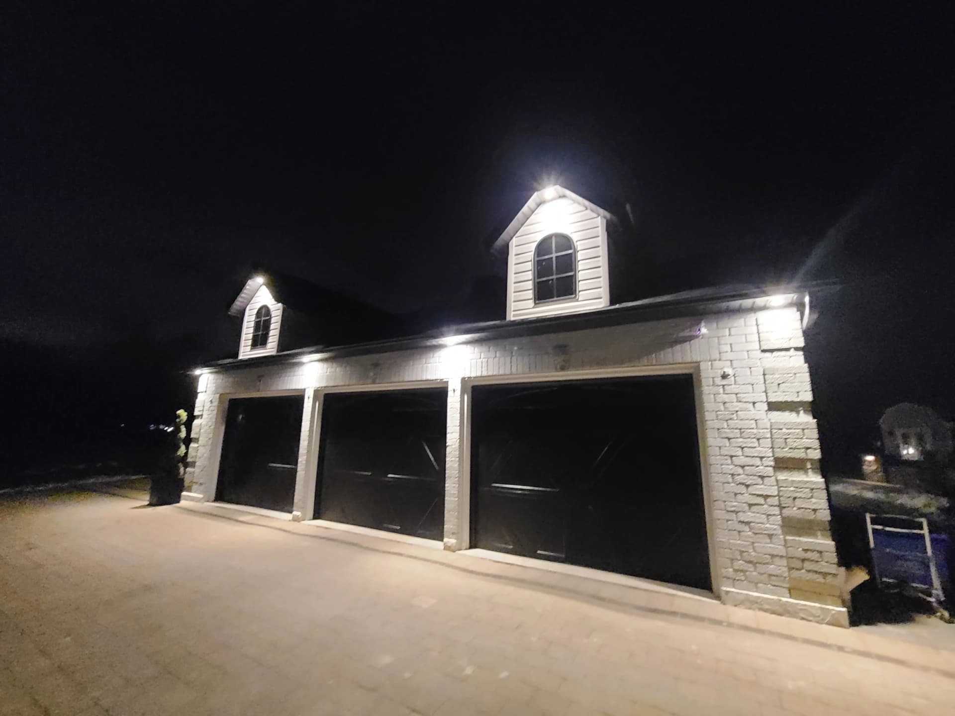 Night photograph of the detached garage at 10 Fielding Court with three dark garage doors, two dormer windows and illuminated stone walls