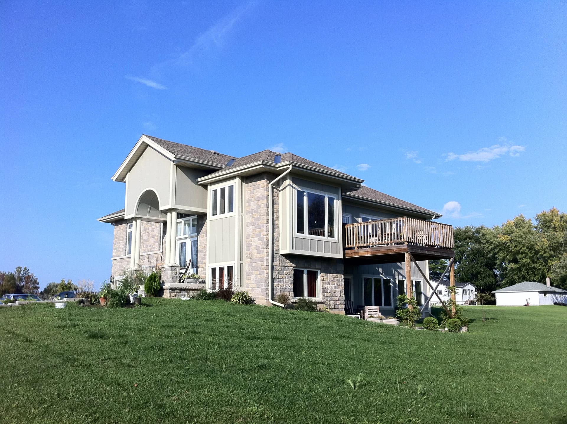 Side photograph of the Regional Road 24 residence showing stone walls, tall windows, the raised timber deck and a sloping lawn