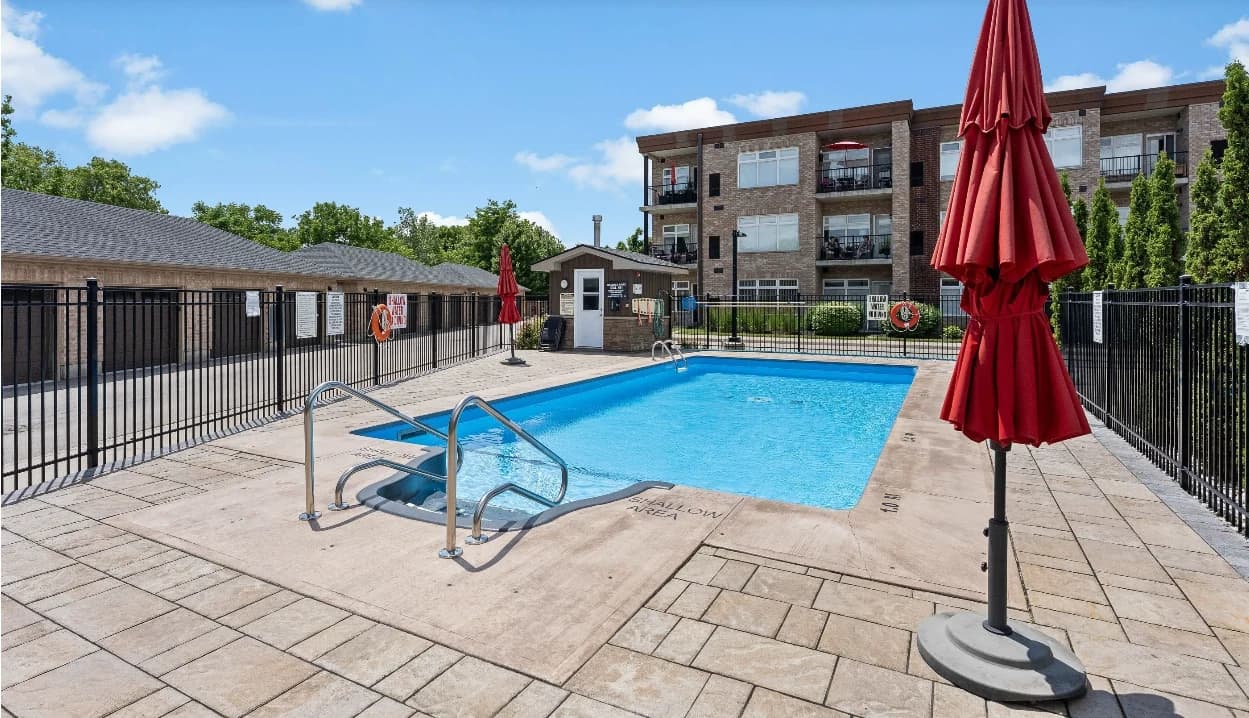 Outdoor swimming pool with entry handrails, a paved terrace and red umbrellas beside Olympia's brick condominium building