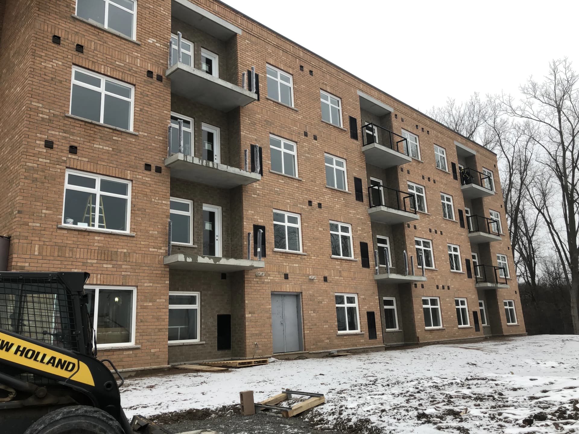 Supplied winter photograph along the balcony elevation at 5629 Swayze Drive showing brick walls, dark railings, ground-floor openings and a construction vehicle